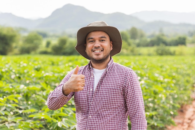 Happy farmer with hat standing showing thumb up and smiling in plantation field at sunset.