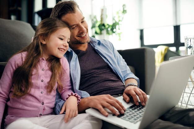 Happy father and daughter sitting close to each other while using computer and surfing the internet at home