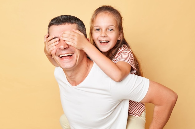 Happy father and daughter wearing casual tshirts standing isolated over beige background daddy caring child on his back kid covering dad eyes and laughing