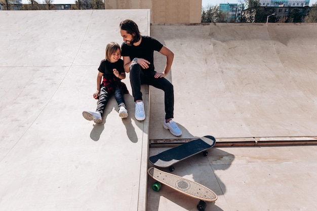 Happy father and his son dressed in the casual clothes are sitting and laughing at the slide in a skate park next to the skateboards at the sunny day .