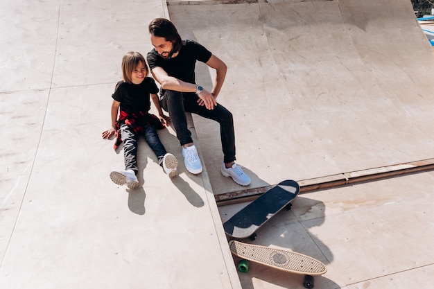 Happy father and his son dressed in the casual clothes are sitting and laughing at the slide in a skate park next to the skateboards at the sunny day .