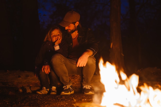 Happy father and his son warm themselves by the fire sitting in an embrace on logs in a hike in the forest at the night. .