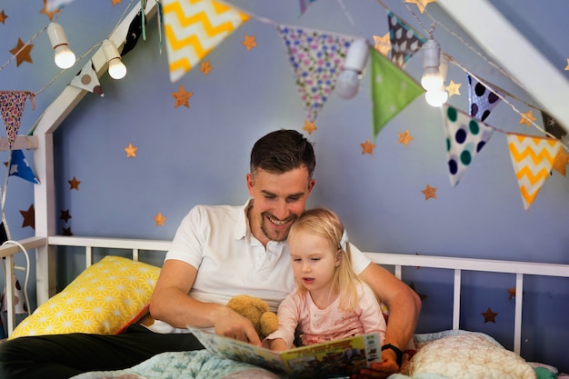 Happy father reading book with his little girl while sitting on bed before bedtime together