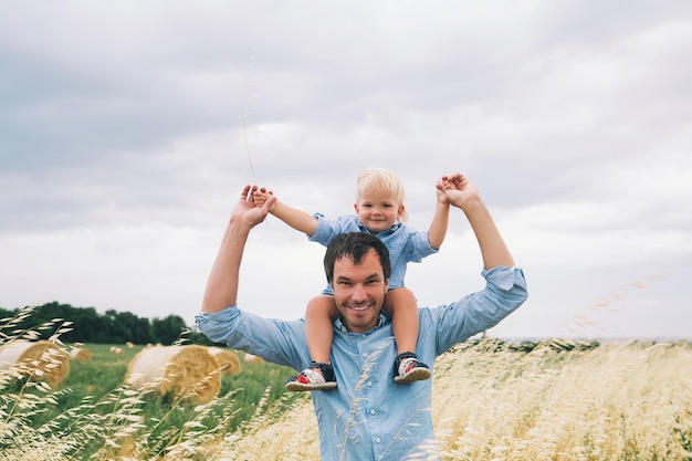 Happy father and son Family concept Man and his little cute kid boy having fun on wheat field with haystacks at summer day outdoors Dad carrying child on his back with raised arms up on nature