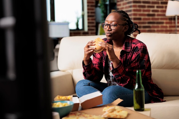 Happy female adult eating burger from fast food delivery takeout, watching movie on tv channel program. Serving takeaway meal to enjoy leisure activity with film on television.