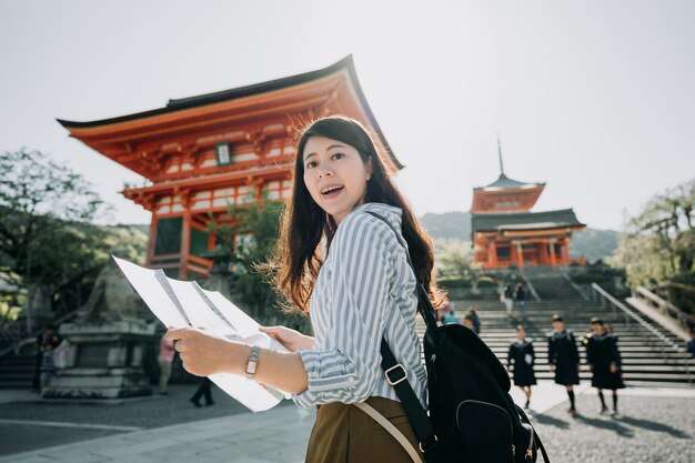 Happy female asian tourist sightseeing kyoto city famous tourism attraction with map. young girl visitor holding guided paper visit kiyomizu temple on sunny day. japanese high school students in back