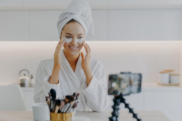 Happy female beauty blogger records video for her blog while applying beauty moisturising patches under eyes wears wrapped towel on head and domestic robe shares opinion about cosmetic product