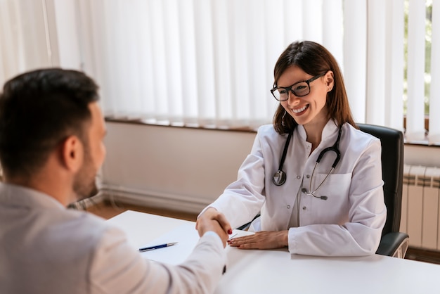 Happy female brunette doctor at medical office with patient shaking hands.