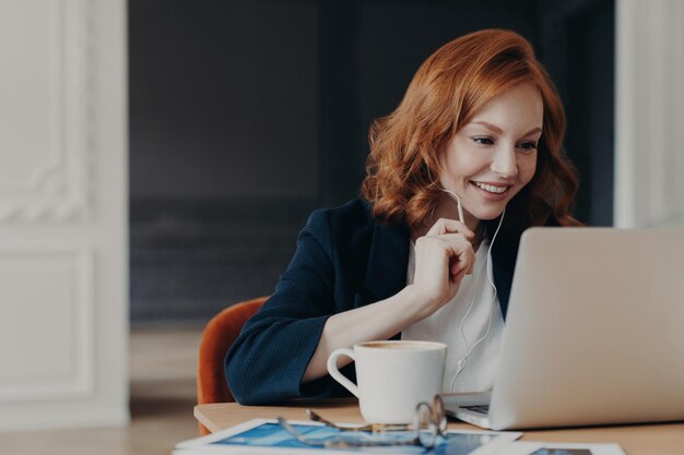 Happy female entrepreneur has online conference uses laptop with wireless internet and earphones at home or coworking space