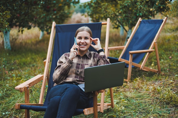 Happy female freelancer working on digital netbook relaxing in garden chair and smiling at camera woman with laptop posing in park during remote work