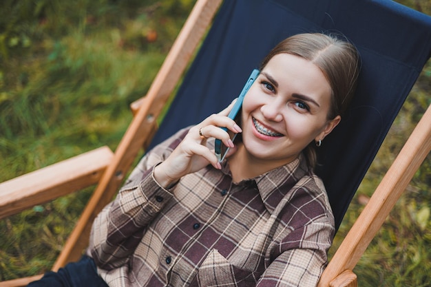 Happy female freelancer working on digital netbook relaxing in garden chair and smiling at camera woman with laptop posing in park during remote work