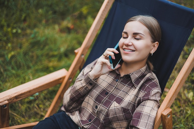 Happy female freelancer working on digital netbook relaxing in garden chair and smiling at camera woman with laptop posing in park during remote work