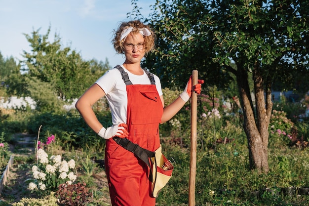 A happy female gardener in gloves and an apron stands against the background of a flower bed in a home garden Gardening and floriculture Flower care