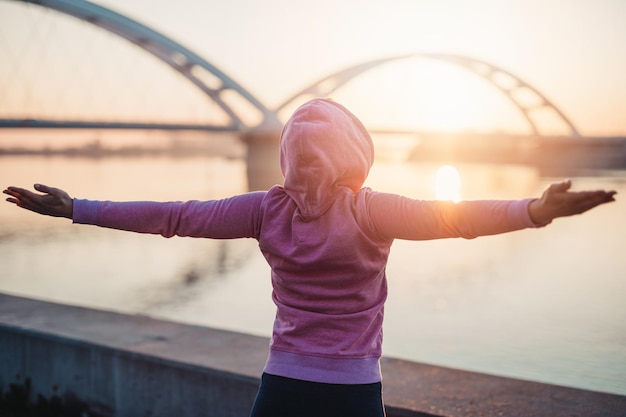 Happy female jogger standing and enjoying in beautiful river sunrise. Winning gesticulation with hands up. View from the back.