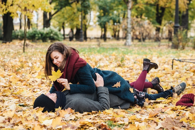 Happy female and male enjoy spending time together, lie on ground covered with yellow leaves