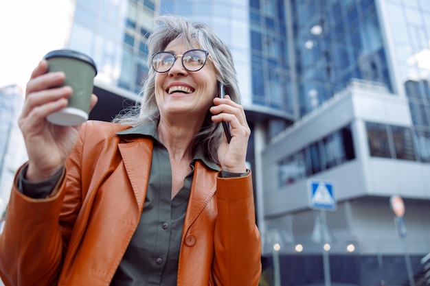 Happy female pensioner with cup of drink n talks on mobile phone on modern city street