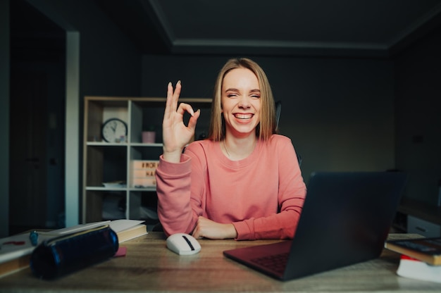 Happy female student sitting at home at desk with laptop and books and posing at camera