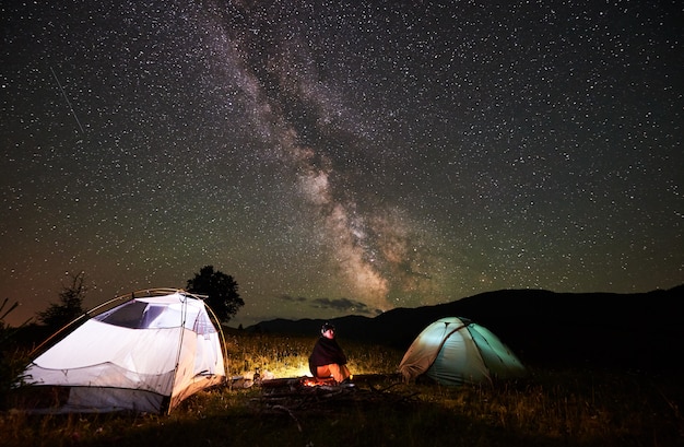 Happy female traveller enjoying incredible beautiful starry sky and Milky way at night camping in the mountains. Woman sitting on a log beside campfire and two illuminated tents.