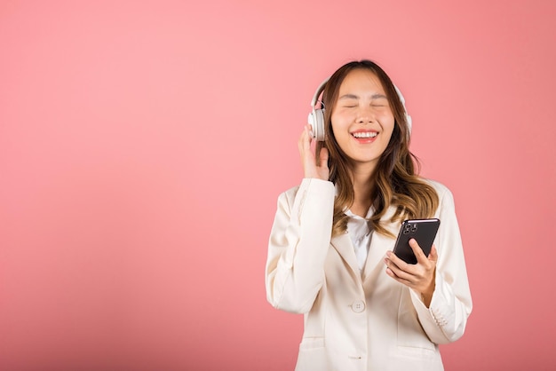 Happy female using mobile phone listens loud song, Portrait of Asian beautiful young woman has fun close eyes listening music with headphones in smartphone app, studio shot isolated on pink background