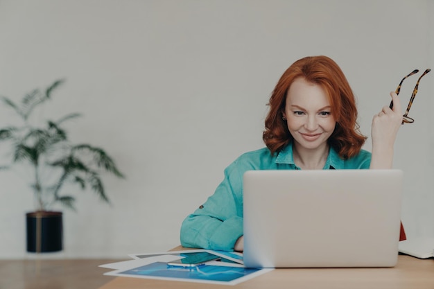 Happy financial analyst beautifully poised conducts economic research consults clients online finishing tasks holding spectacles and working diligently on her laptop amidst documents at her desk