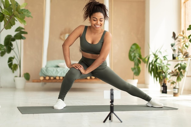 Happy fit black lady in sportswear doing side lunge exercise and looking at smartphone on tripod training at home