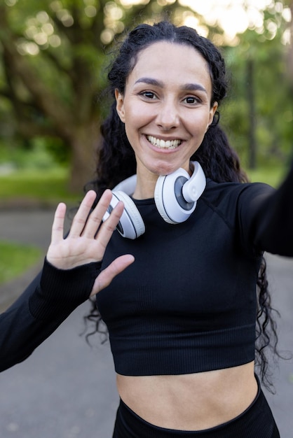 A happy fit young woman is seen waving at the camera wearing headphones around her neck and workout