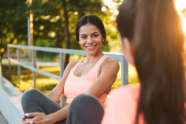 happy fitness women in sportswear smiling and talking together while sitting in green park