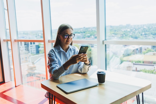 Happy freelancer girl in classic glasses looking at phone and smiling while sitting in modern coworking space carefree millennial woman in glasses enjoying leisure time for communication