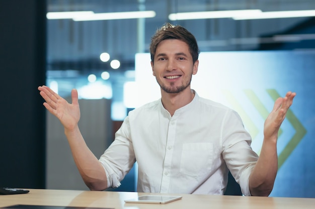 A happy and friendly young man in the office is sitting at a table showing with his hands that he is glad to see you Advertises the product