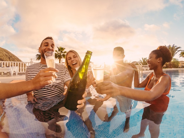 Happy friends cheering with champagne in pool party at sunset