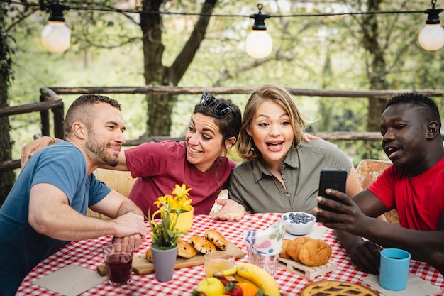 Happy friends having breakfast in the backyard young influencer using smartphone to create content on social media with photos of food multiracial couples having fun at picnic outside