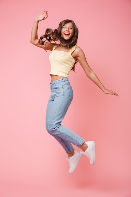 Happy full length photo of cheerful woman with long brown hair rejoicing and jumping, isolated over pink background