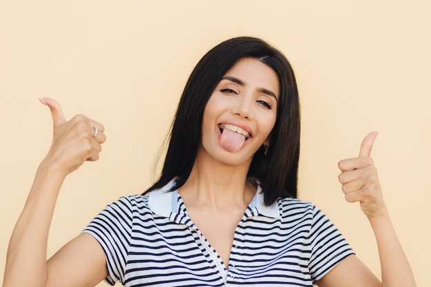 Happy funny young female with pleasant appearance gestures with both hands shows okay sign and tongue blinks with eye has funny look isolated over beige background People and positiveness