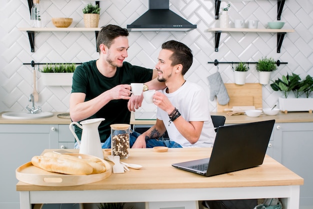 Happy gay couple drinking coffee and having fun together. Everyday morning routine of gay couple life in kitchen at home. Gay concept, happy life