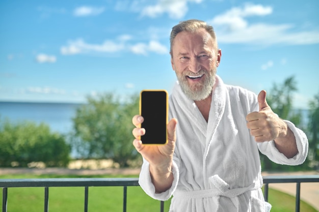 Happy gentleman in the white bathrobe demonstrating his smartphone