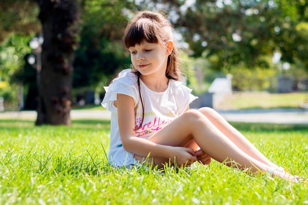 Happy girl of 8 years old in a white dress and long dark hair sits on a green lawn