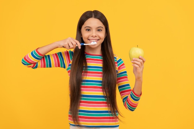 Happy girl child brush teeth with toothbrush holding apple yellow background, healthy teeth.