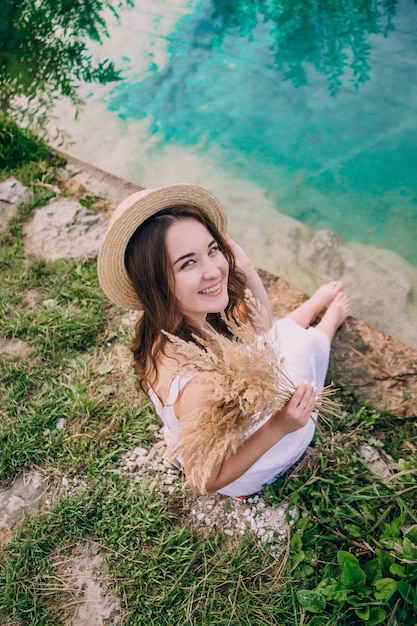 Happy girl in a dress sitting on the shore of a blue lake. Girl in a straw hat with a bouquet near a small lake.