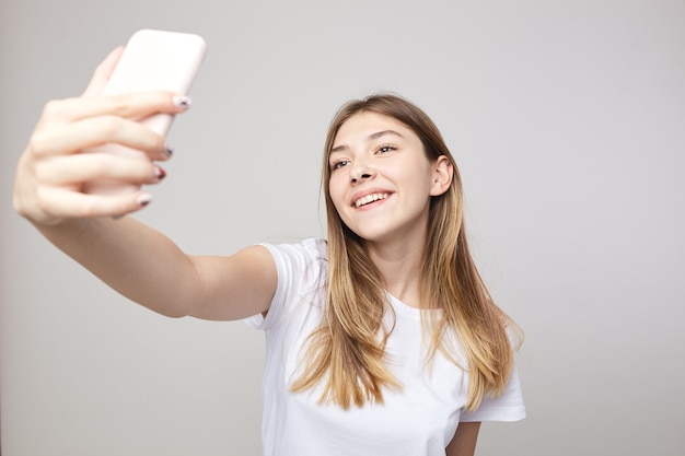 Happy girl dressed in a white tshirt makes selfie on a white background in the studio