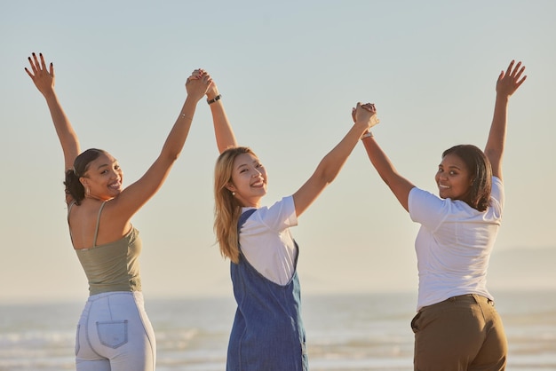 Happy girl friends and holding hands at a beach with women on a holiday freedom and travel Portrait of people with arms raised to show excited adventure by the ocean and sea together by water