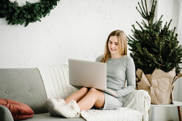 Happy girl greeting friends or family with New year in video chat on laptop Young woman using laptop at home office near decorated pine tree at home with Christmas interior with copy space