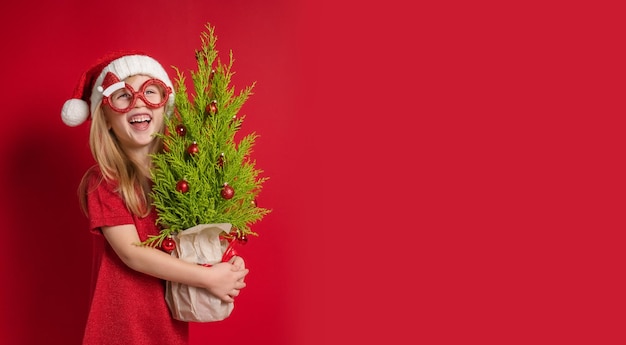 Happy girl holding a Christmas tree with decorations in her hands Red background with copy space