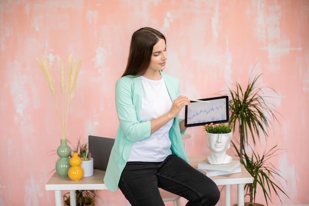 Happy girl holding tablet with financial graphic and showing at it in colorful office