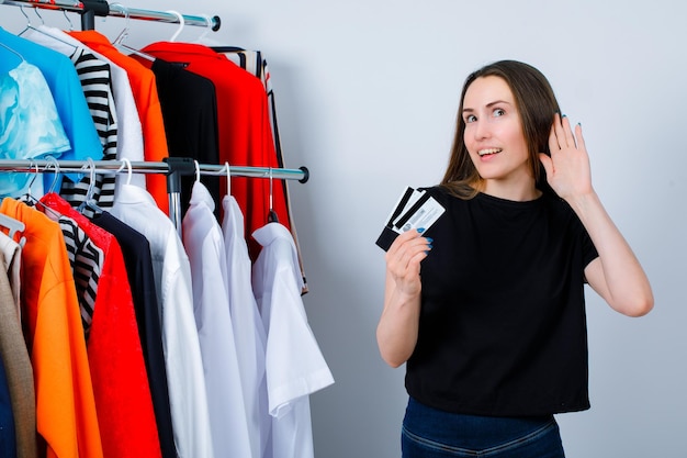 Happy girl is holding credit cards and holding other hand near ear on clothes background