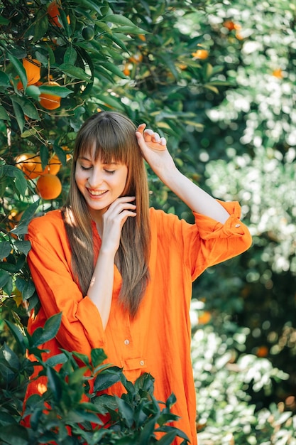 Happy girl in orange dress is posing to camera by looking down and holding up her hands in orange garden