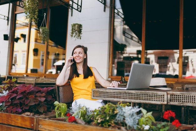 Happy girl in outdoors street cafe sitting at table with laptop pc computer, talking on mobile phone, conducting pleasant conversation, in restaurant during free time