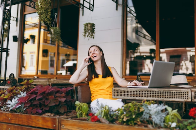 Happy girl in outdoors street cafe sitting at table with laptop pc computer, talking on mobile phone, conducting pleasant conversation, in restaurant during free time