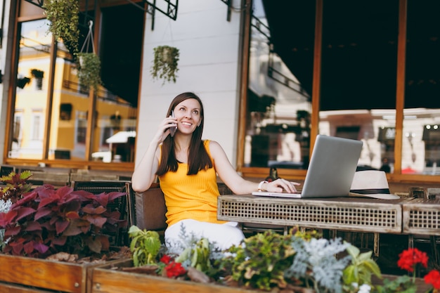 Happy girl in outdoors street cafe sitting at table with laptop pc computer, talking on mobile phone, conducting pleasant conversation, in restaurant during free time