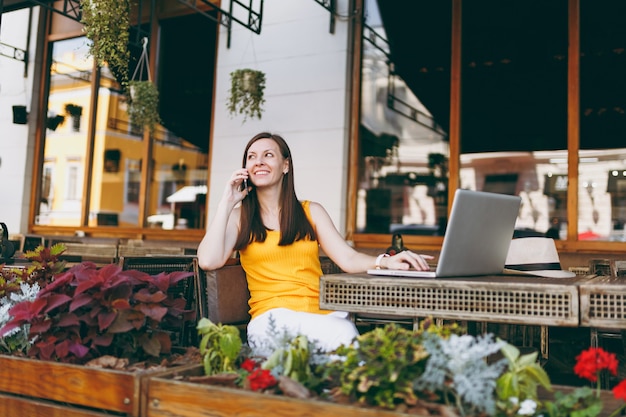 Happy girl in outdoors street cafe sitting at table with laptop pc computer, talking on mobile phone, conducting pleasant conversation, in restaurant during free time