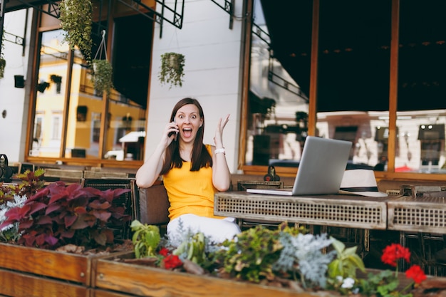 Happy girl in outdoors street cafe sitting at table with laptop pc computer, talking on mobile phone, conducting pleasant conversation, in restaurant during free time